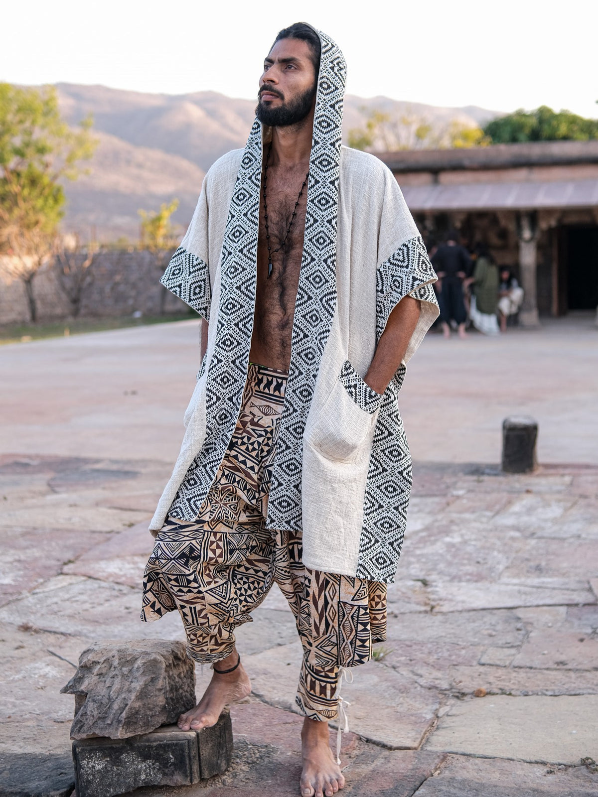A man wearing a hooded, light-colored Neer poncho with a black-and-white geometric pattern over patterned, loose-fitting trousers.
