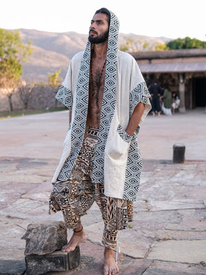 A man wearing a hooded, light-colored Neer poncho with a black-and-white geometric pattern over patterned, loose-fitting trousers.