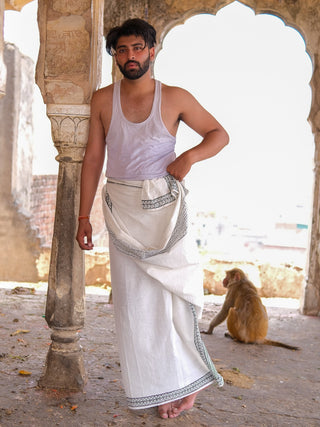 A man in a Nilaya Dhoti and white vest stands by a stone pillar in an arched building, sunlight streaming in as a monkey sits behind him, adding to the serene atmosphere.