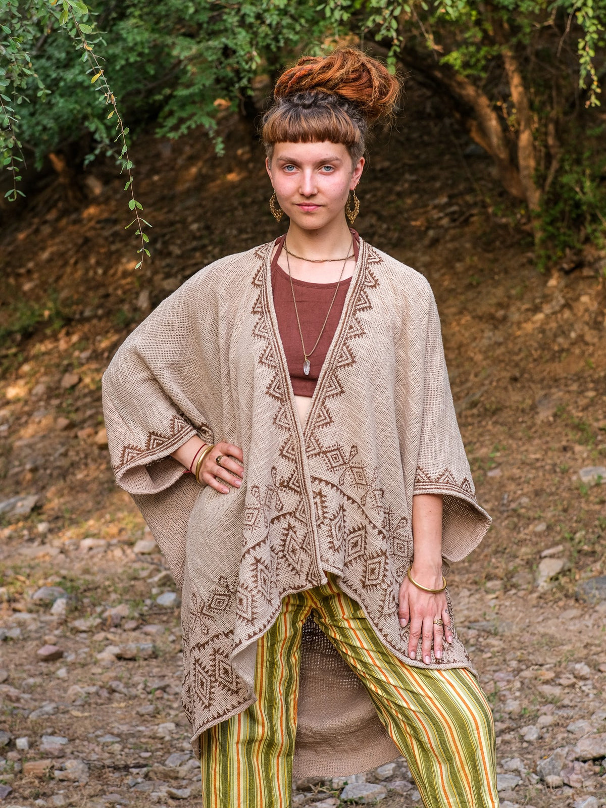 A woman stands outdoors wearing a brown top, green-yellow striped pants, and the elegant Miraya Shrug—a tan patterned kimono-style layer reminiscent of a block print. She has her hair in a top bun and accessorizes with layered jewelry, surrounded by trees and rocky ground.