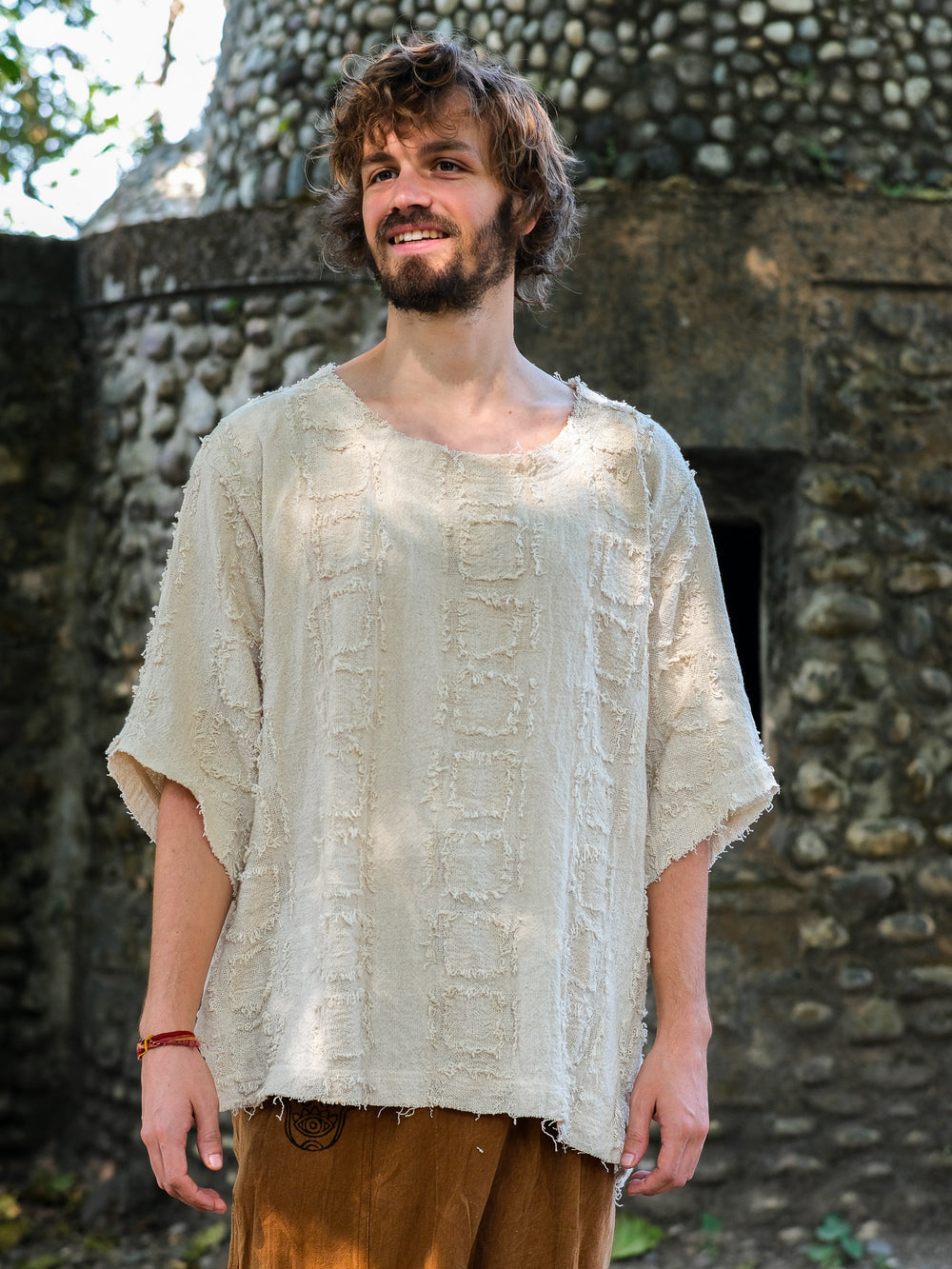 A man with wavy brown hair and a beard stands outdoors, smiling. He wears the Lennon T-shirt in Natural (100% cotton, baggy fit) and brown pants for a relaxed bohemian look. Behind him is a stone wall with a small dark opening.