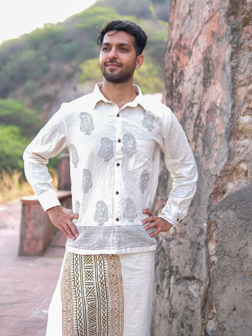 A man stands outdoors, smiling slightly with his hands on his hips, wearing a cream-colored Shakti Shirt featuring printed patterns, set against a backdrop of a stone wall and greenery.