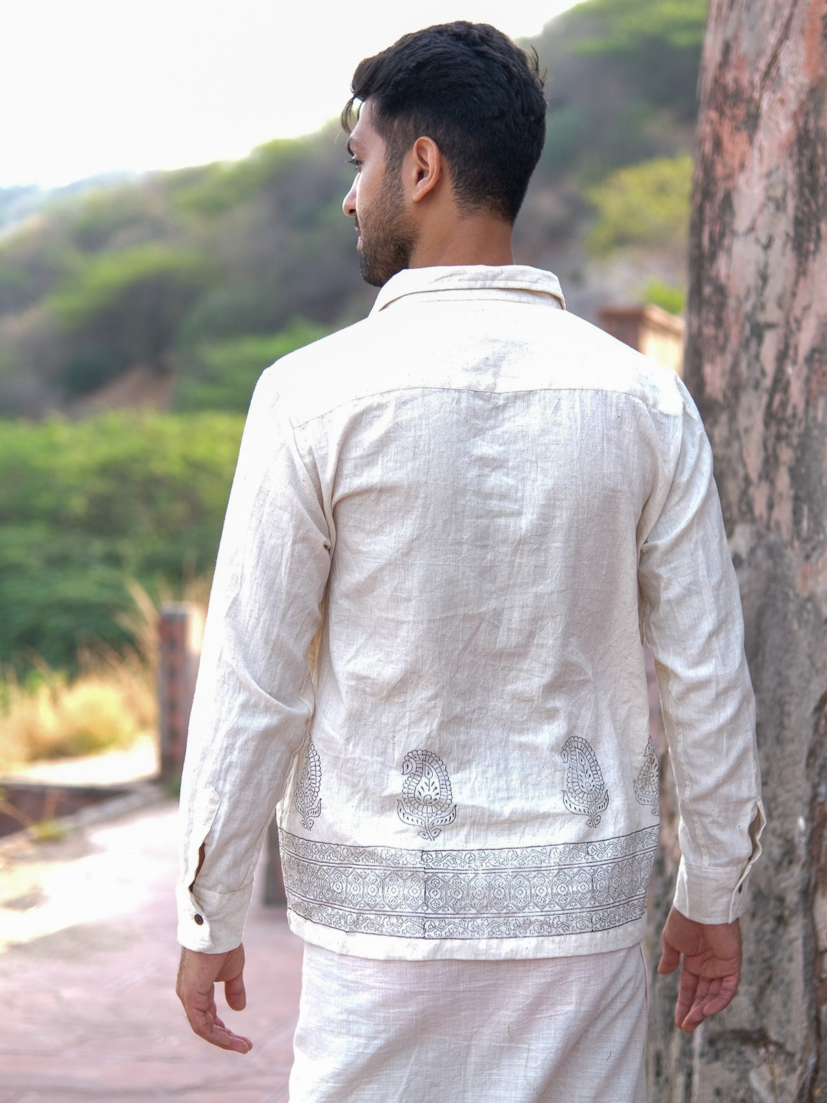 A man stands outdoors with his back turned, wearing the light-colored Shakti Shirt adorned with intricate silver patterns along the hem. Lush green foliage and stone structures form the backdrop.