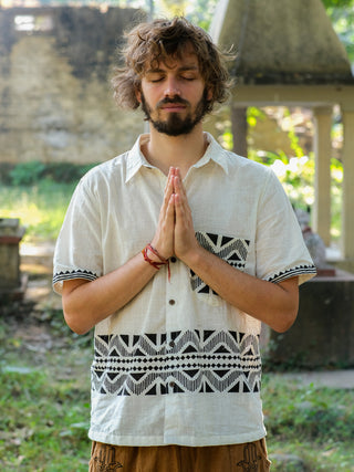 A closed-eyed person stands meditatively with hands in prayer, wearing a Beatle shirt with Rajasthan handblock print for a vibrant, eco-friendly look. Theyre outdoors amidst blurred greenery and a stone structure in the background.