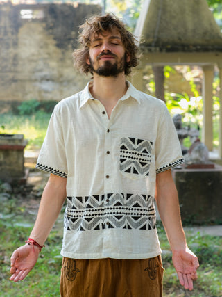 A curly-haired person outdoors meditates or relaxes with closed eyes and open arms in front of greenery and a stone structure, wearing a Rajasthan handblock Beatle shirt and brown pants.