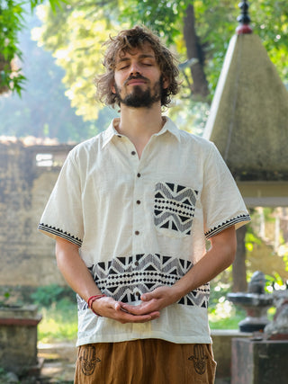 With eyes closed and curly hair, a person stands serenely outdoors wearing a patterned Beatle shirt reminiscent of Rajasthan handblock print, paired with brown pants. Their hands gently clasped in front, they are framed by trees and a stone structure in the background.