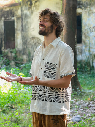 A curly-haired, bearded man smiles and gestures, wearing a Beatle shirt with Rajasthan handblock print and brown pants. He stands outdoors amidst grass, trees, and an old building.