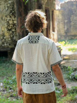 A person with wavy brown hair is outdoors in a light Beatle shirt with Rajasthan handblock geometric black patterns, set against an old stone building on green grass, sunlight filtering through trees.