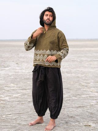 Barefoot on a barren landscape, a person dons a Tribal Hoodie featuring white geometric patterns on green fabric, paired with loose black pants. They stand looking thoughtfully into the distance, hand near chin.