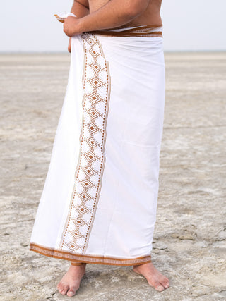 In a dry, barren landscape, a person wears the Kangura Dhoti made of 100% cotton. The traditional white fabric, with a brown and gold geometric border, is elegantly draped and tied at the waist, while their feet connect to the earth below.