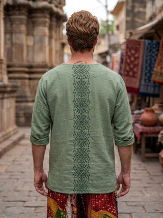 A man with short, light brown hair stands on a stone street, facing away. He wears the Noma Shipibo Kurta in light green—crafted from breathable fabric and featuring a vertical geometric Shipibo hand block print on the back—paired with colorful patterned pants. Fabric stalls and stone buildings create the backdrop.