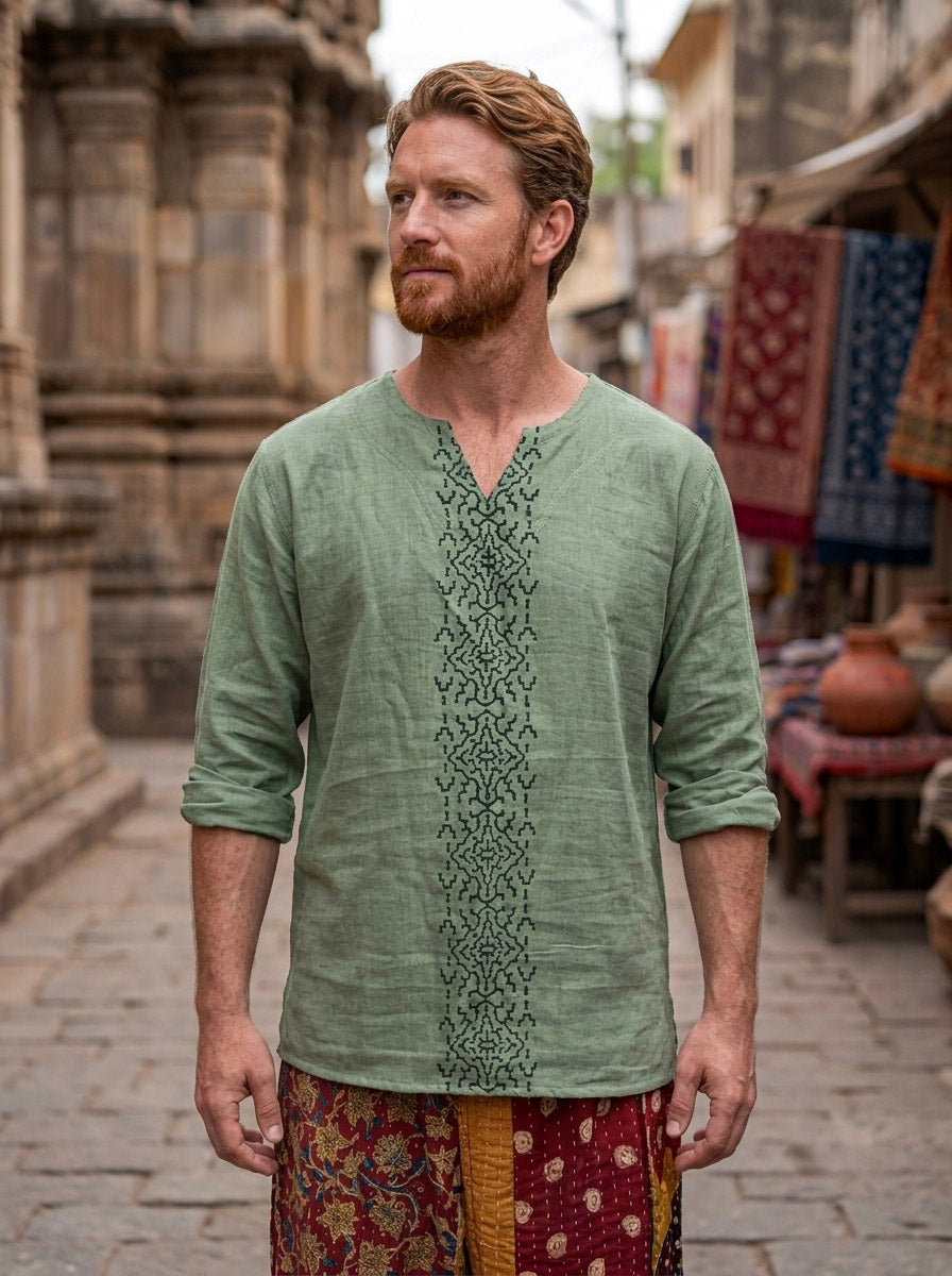 A man with red hair and a beard stands in a marketplace wearing the mint green Noma Shipibo Kurta, crafted from lightweight double gauze cotton and adorned with subtle hand block print embroidery, paired with patterned pants. Stone buildings and stalls draped in colorful fabrics set a vibrant backdrop for the scene.