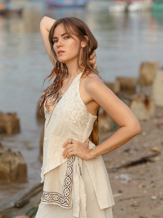 Confidently standing by the waterfront, a person with long brown hair wears feather earrings and the HWC Triveni Top, a sleeveless white dress featuring a v-neck design. The scene is serenely set with water, boats, and concrete blocks in the background.