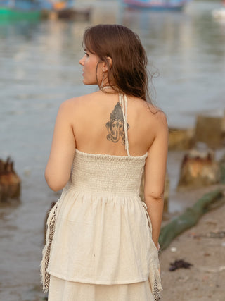 A person with long brown hair, wearing the HWC Triveni Top with an intricate block print, stands by the water, showcasing an elephant tattoo on their upper back. The backdrop features a blurred shoreline and boats drifting peacefully in the distance.