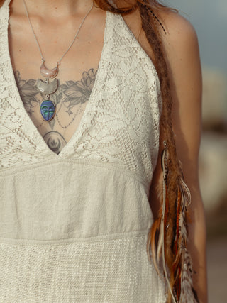 Wearing a Triveni Top by HWC, a person with visible tattoos and long braided hair embellished with adornments sports a pendant necklace featuring crescent moon and face motifs, set against a softly blurred background.