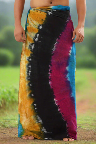 A person is outdoors wearing a Tiedye Dhoti in black, yellow, blue, and pink hues. The blurred greenery enhances the bohemian charm of the scene, as they stand barefoot with their hands at their sides.