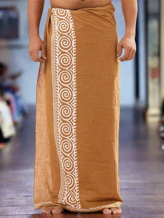 A person in a brown and white Tribal Dhoti, adorned with intricate spirals, stands barefoot. The Rajasthani hand block-print fabric drapes elegantly around the waist against a blurred background, accentuating its traditional Indian style.