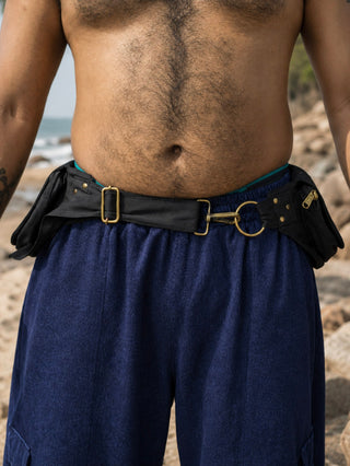A person wearing blue pants and the Mystic Waist Belt with gold buckles stands on a rocky beach, their bare torso partially visible.