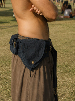 A person standing outdoors, shirtless with arms crossed, is wearing loose brown pants and the Black Rustic Festival Bag around their waist. The scene captures a bohemian style atmosphere, with grass and people visible in the background.