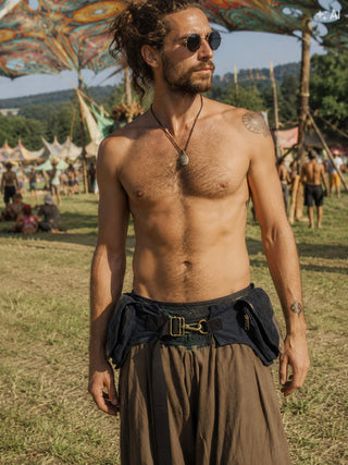 A shirtless man with sunglasses and a pendant stands outdoors at a festival, wearing loose brown pants and accessorizing his look with the Rustic Festival Bag—a utility belt with pouches. Colorful tents and people in the background enhance the bohemian style atmosphere.