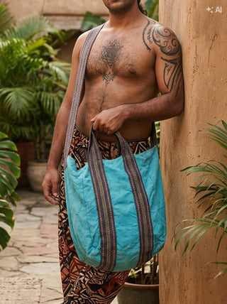 A shirtless person with tattoos stands outdoors next to plants and a textured tan wall, holding the Anoop Tote Bag—an oversized, bohemian-style tote made of turquoise fabric with patterned straps.