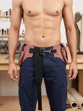 A shirtless person stands in a workshop wearing blue cargo pants and a Rustic Festival Bag—a brown utility belt featuring zippered pouches and a black strap. Tools and materials are visible on the workbench behind them.