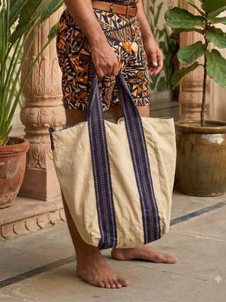 A person in patterned shorts stands barefoot on a stone floor, holding the Anoop Tote Bag—a large beige cotton canvas tote with dark blue handles. There are potted plants and carved columns in the background.