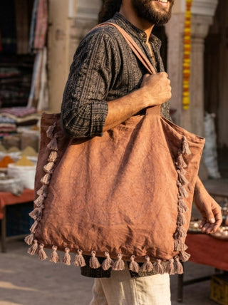 A person wearing a dark patterned shirt carries the Aatman Tassel Tote Bag, featuring hand block printed bohemian motifs and tassels along the edges, while walking outdoors in a market setting.