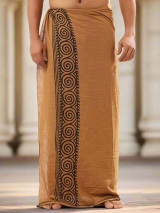 A person stands barefoot on a stone surface, wrapped in the brown and black Tribal Dhoti, featuring a traditional Rajasthani spiral pattern border that showcases the intricate hand block-print style.