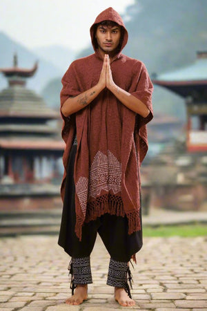 A barefoot person stands on stone pavement, hands pressed in prayer, wearing a hooded brown poncho draped with the Swing Shawl and patterned black pants, with traditional architectural structures visible in the blurred background.