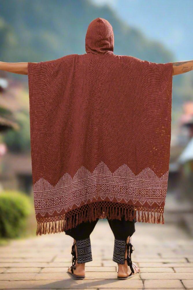 A person stands outdoors with arms outstretched, wearing the Swing Shawl—a hooded, patterned brown poncho with fringe at the bottom that captures the charm of a traditional shawl—and black pants featuring decorative cuffs. The background is blurred.