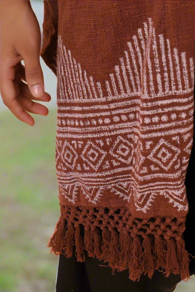 A hand is next to the Swing Shawl, a brown woven traditional shawl with white geometric patterns and tassels at the bottom. The background is blurred green and light brown.