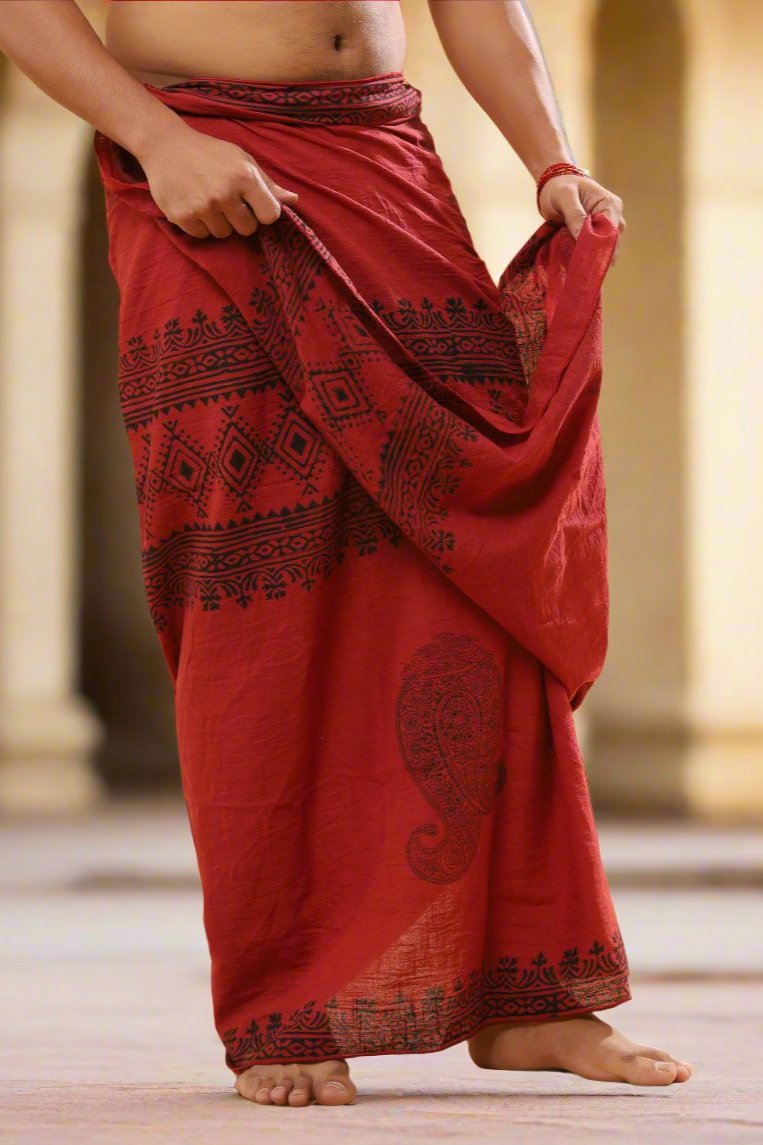 Model holding the waist folds of a red Vasuki handblock print cotton dhoti, warm stone-pillared backdrop
