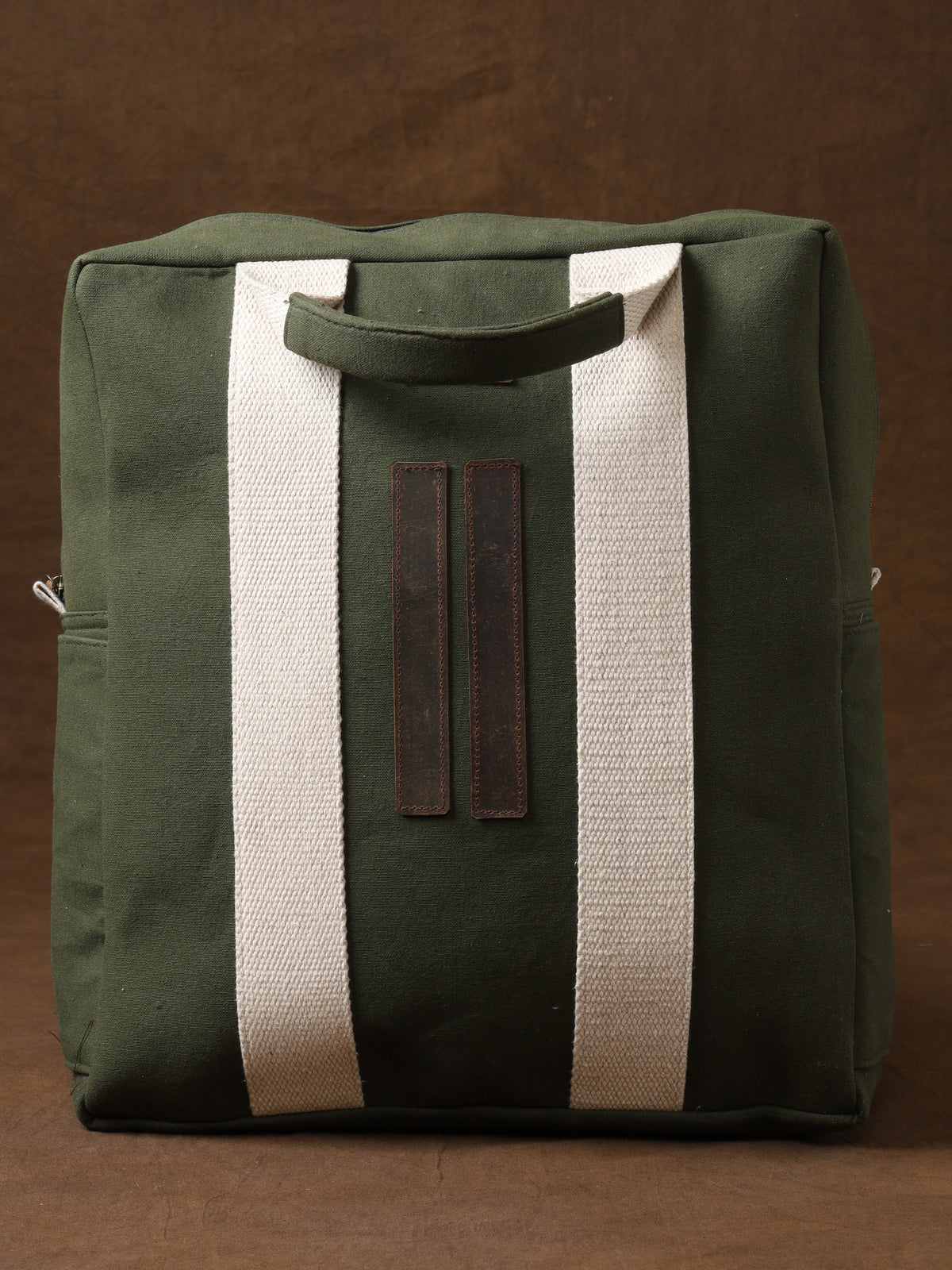Kelly Back Pack in green canvas with wide white straps and brown leather accents, displayed against a brown background. This square-shaped bag features top handles—an ideal adventure companion for those who value both functionality and style in their travel gear.