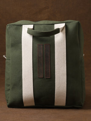 Kelly Back Pack in green canvas with wide white straps and brown leather accents, displayed against a brown background. This square-shaped bag features top handles—an ideal adventure companion for those who value both functionality and style in their travel gear.