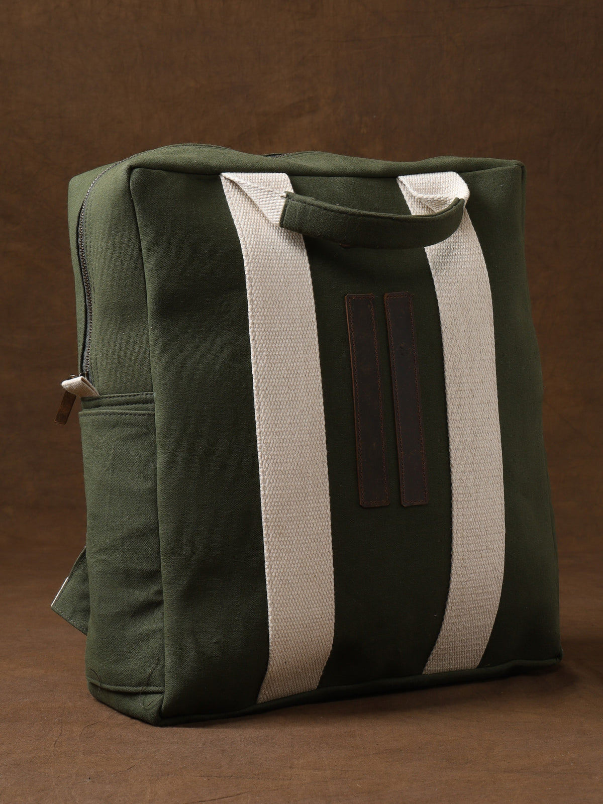 The Kelly Back Pack—a green canvas tote with white straps and a zipper—stands upright against a brown background, making it your ideal adventure companion for everyday outings.