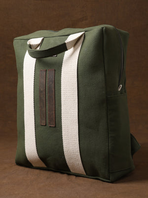 Introducing the Kelly Back Pack: a green canvas tote bag featuring two sturdy white handles and sleek vertical brown leather accents, showcased upright against a rich brown background—your ideal adventure companion, merging practicality and elegance in true Kelly Back Pack style.
