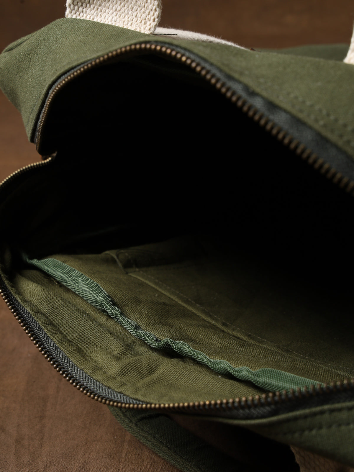 The Kelly Back Pack, an open olive green fabric bag featuring a visible zipper, white handles, and a roomy inner compartment, is displayed on a brown surface—making it the perfect companion for any adventure.