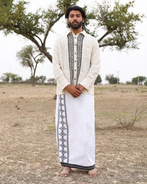 A man stands barefoot outdoors on dry grass, wearing the cream-colored Acharya Shirt Full-sleeve—crafted from organic cotton and adorned with black patterns—and a white traditional wrap featuring matching black designs. Sparse trees and a cloudy sky are in the background.