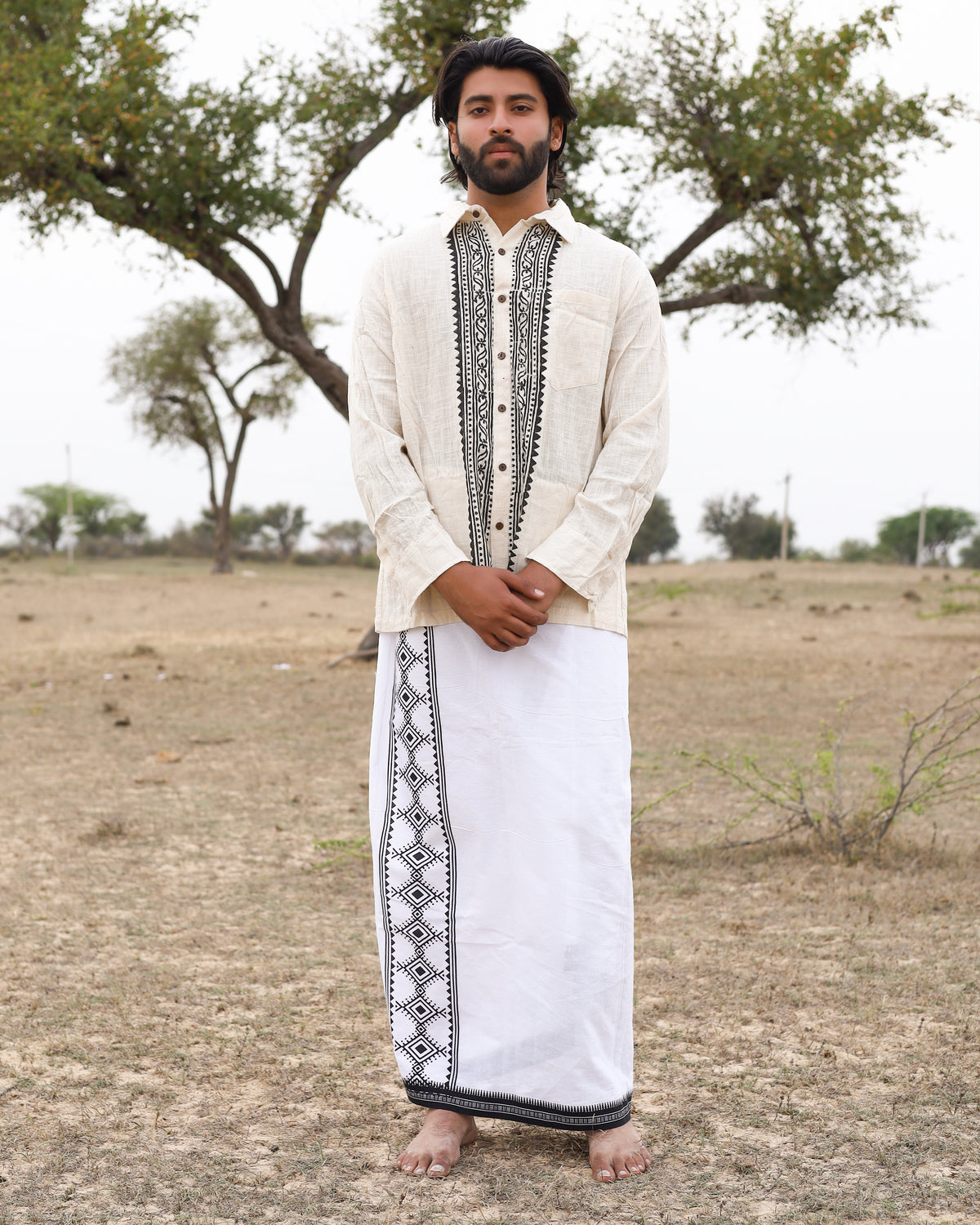 A man stands barefoot outdoors on dry grass, wearing the cream-colored Acharya Shirt Full-sleeve—crafted from organic cotton and adorned with black patterns—and a white traditional wrap featuring matching black designs. Sparse trees and a cloudy sky are in the background.