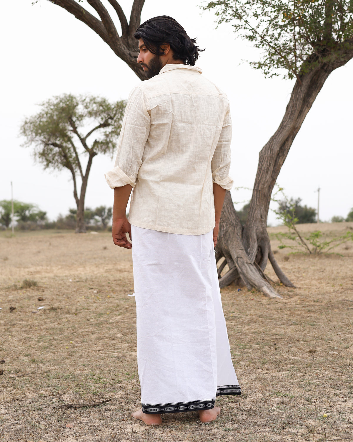 A man with dark hair stands outdoors on dry grass, wearing the Acharya Shirt Full-sleeve in a minimalistic cream tone and a white traditional dhoti with a black border, facing away from the camera. Sparse trees are visible in the background.
