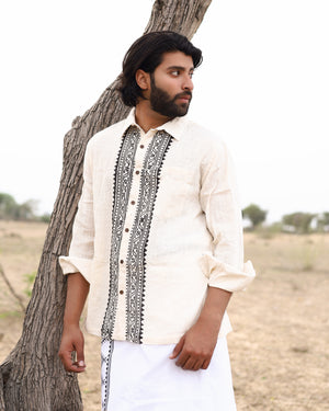 A man with dark hair and a beard stands outdoors near a tree, wearing the Acharya Shirt Full-sleeve in natural cream, crafted from organic cotton. The shirt features intricate black block prints and rolled-up sleeves, paired with a white garment. A serene field dotted with trees is in the background.