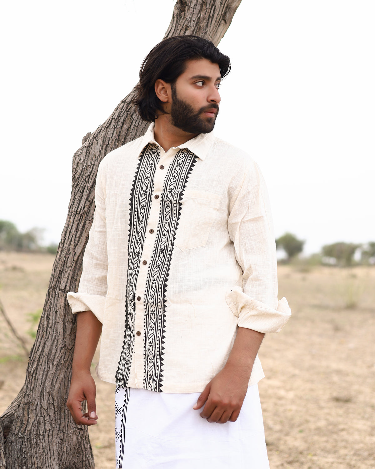 A man with dark hair and a beard stands outdoors near a tree, wearing the Acharya Shirt Full-sleeve in natural cream, crafted from organic cotton. The shirt features intricate black block prints and rolled-up sleeves, paired with a white garment. A serene field dotted with trees is in the background.