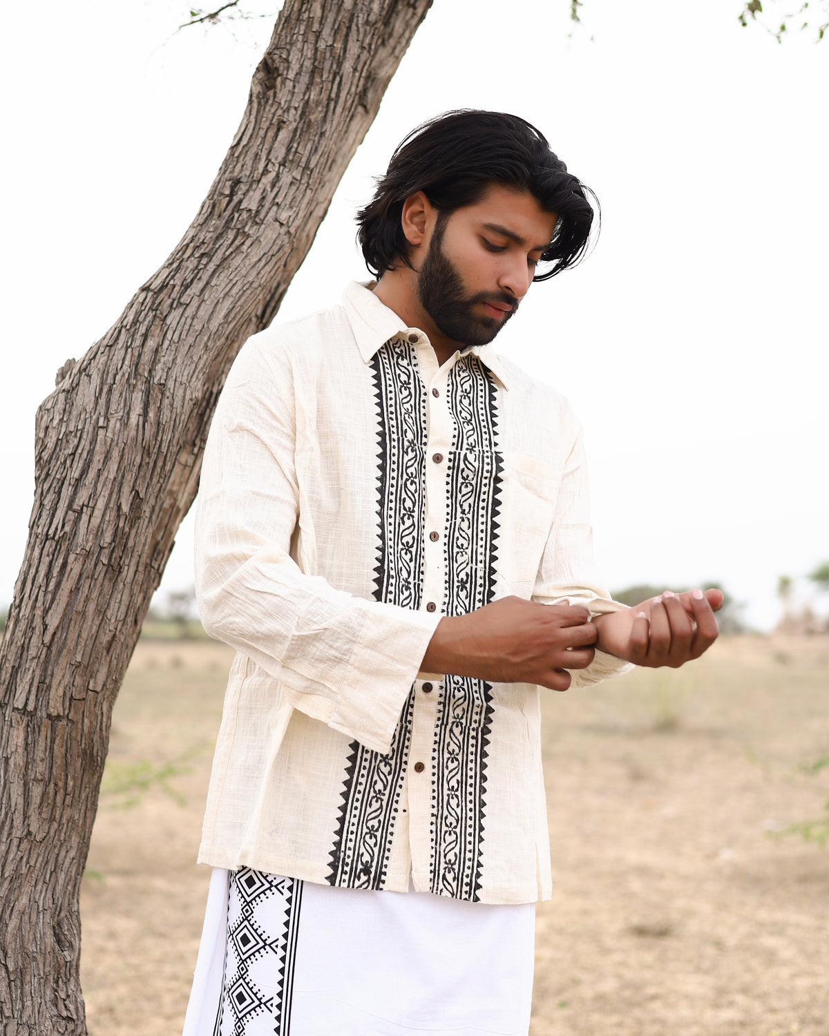 A man with medium-length dark hair and a beard adjusts his sleeve while wearing the Acharya Shirt Full-sleeve, a cream and black embroidered minimalistic design, next to a tree in a grassy field under a cloudy sky.