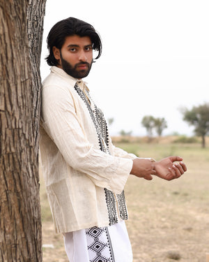 A man with long dark hair and a beard leans against a tree in an open, grassy field. Wearing the Acharya Full-sleeve Shirt with block print designs, his neutral expression meets the cameras gaze amidst trees scattered around.