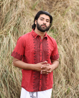 A man with dark hair and a beard stands outdoors before tall dry grass, wearing the eco-friendly, hand-blockprinted red Likhit Shirt and a white cloth around his waist, looking calmly at the camera.