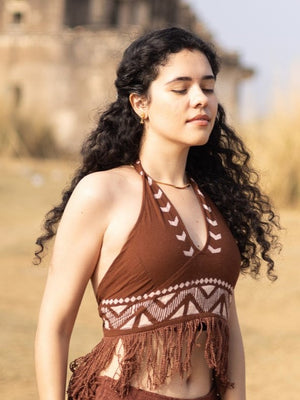 A woman with curly dark hair, wearing the Divine Crop Top featuring fringe details and boho-inspired style, stands outdoors with her eyes closed, radiating a calm and relaxed demeanor against a backdrop of blurred buildings and dry grass.