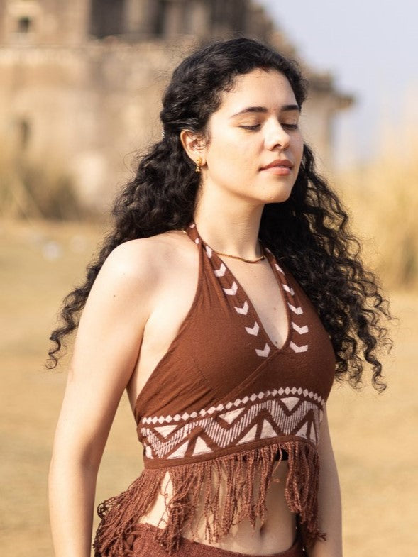 A woman with curly dark hair, wearing the Divine Crop Top featuring fringe details and boho-inspired style, stands outdoors with her eyes closed, radiating a calm and relaxed demeanor against a backdrop of blurred buildings and dry grass.