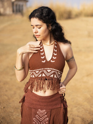 A woman with curly hair stands outdoors wearing the Divine Crop Top—a boho-inspired brown patterned halter with fringe—paired with a matching skirt. She gazes downward, one hand on her shoulder and the other resting on her hip, set against a background of dry grass and blurred buildings that complement the earthy, free-spirited vibe of her block print ensemble.