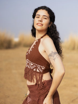 A woman with long curly hair stands outdoors in sunlight, smiling slightly. She is wearing the Divine Crop Top and a matching skirt with a brown block print pattern. A floral tattoo is visible on her upper arm. The background features blurred tall grass.